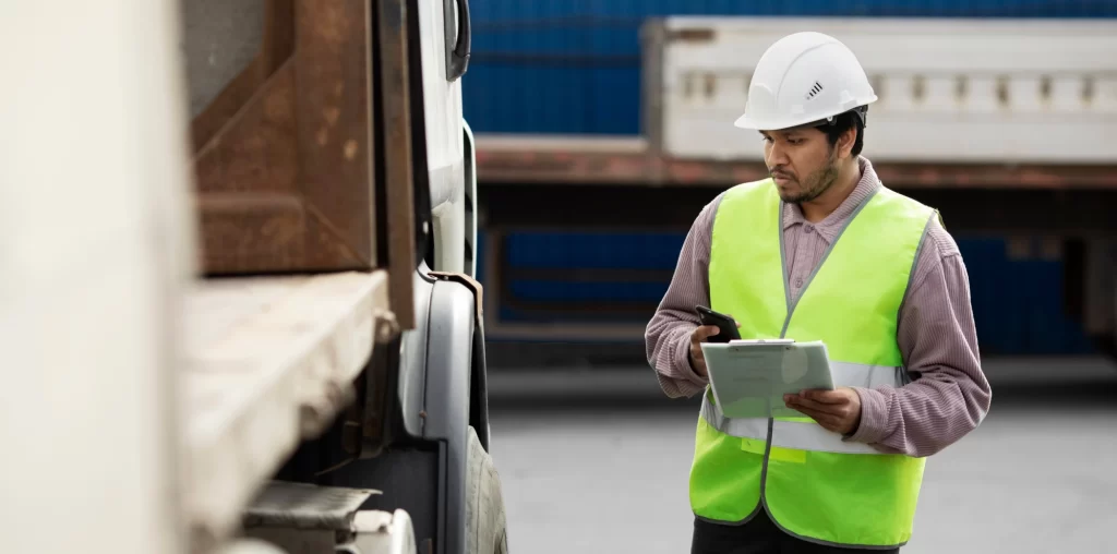 Transport supervisor in safety vest and hard hat checking truck logistics using clipboard and smartphone at loading site.