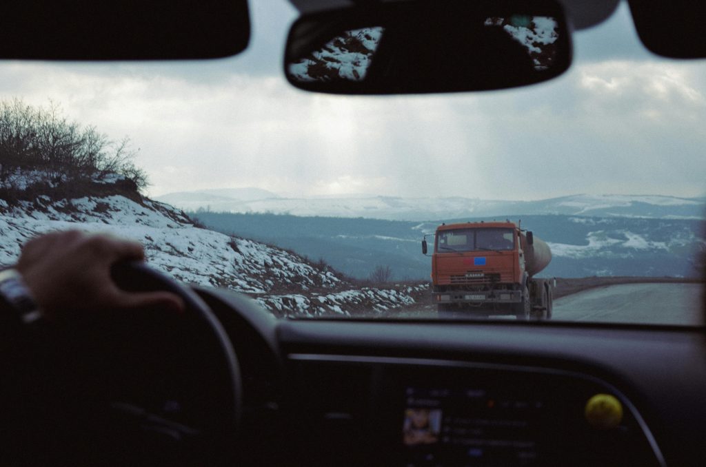 View from inside a car of a cargo truck driving along a winding mountain road with snowy hills in the distance.