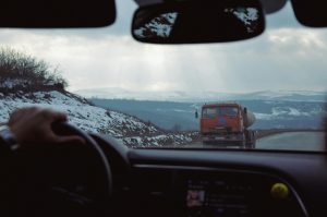 View from inside a car of a cargo truck driving along a winding mountain road with snowy hills in the distance.