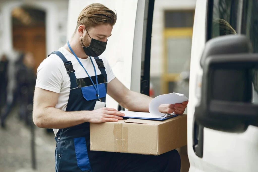 A professional delivery driver wearing a mask checks a clipboard and package to ensure order accuracy, supported by GPS tracking software for on-time delivery.