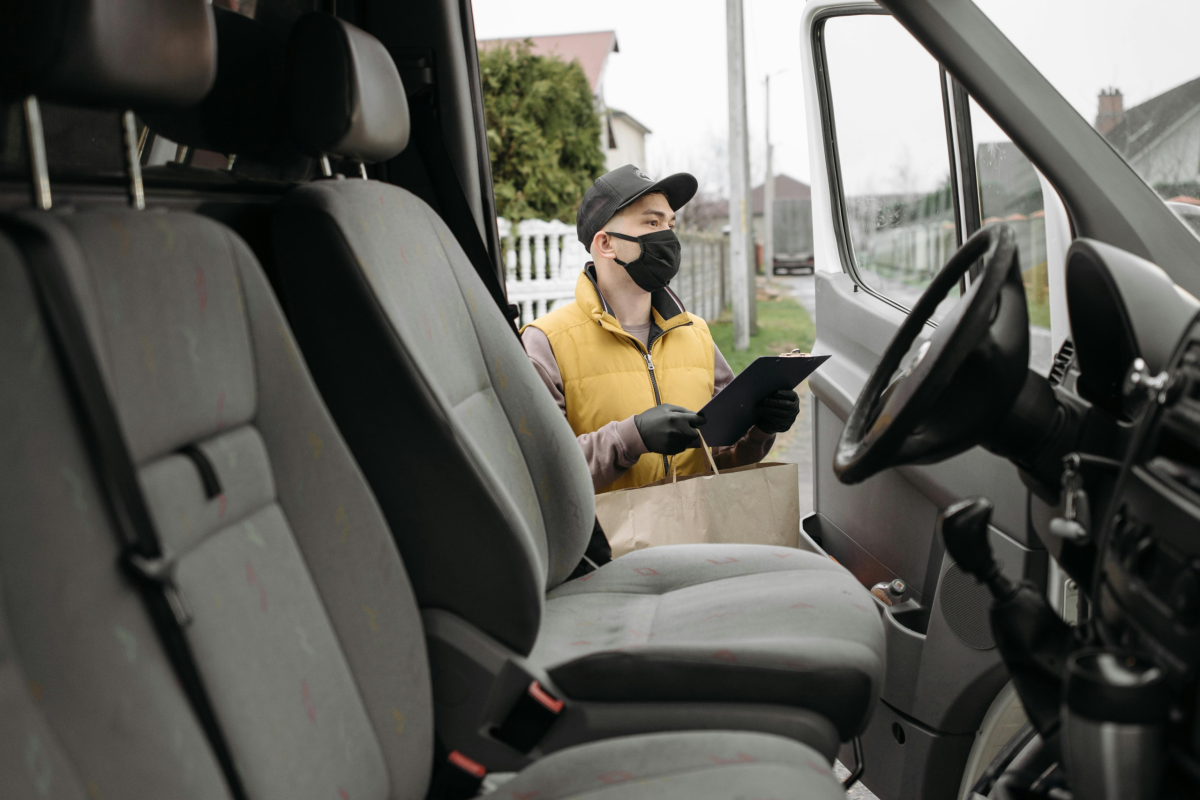 Delivery driver wearing face mask and gloves with clipboard and shopping bag, standing beside a fleet vehicle—showcasing last-mile visibility and real-time tracking with fleet software.